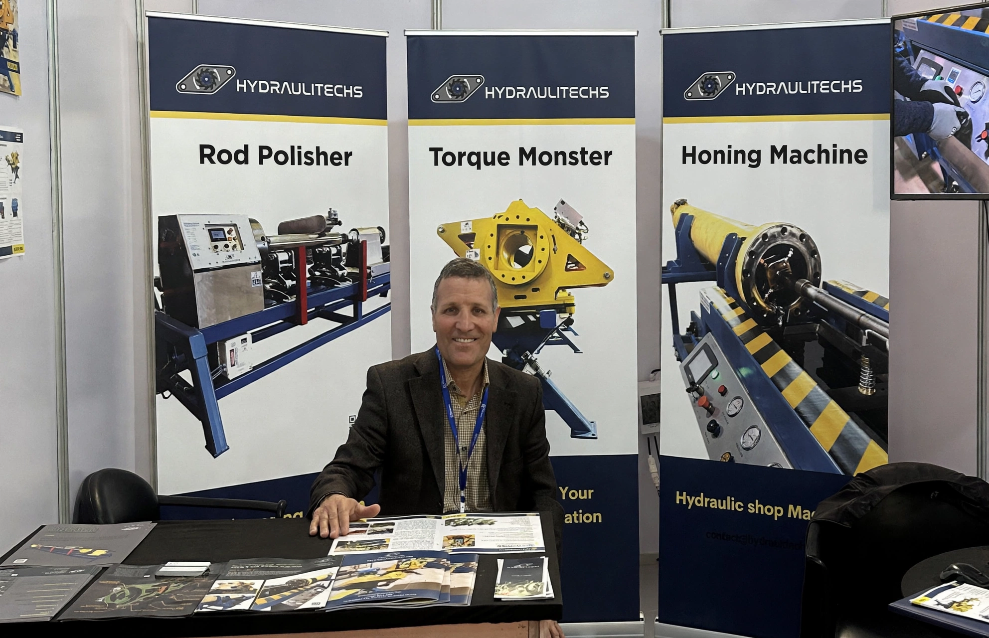 Man Sitting At An Exhibition Booth For Hydraulitechs, With Display Banners Showcasing Rod Polisher, Nutbuster, And Honing Machine Equipment